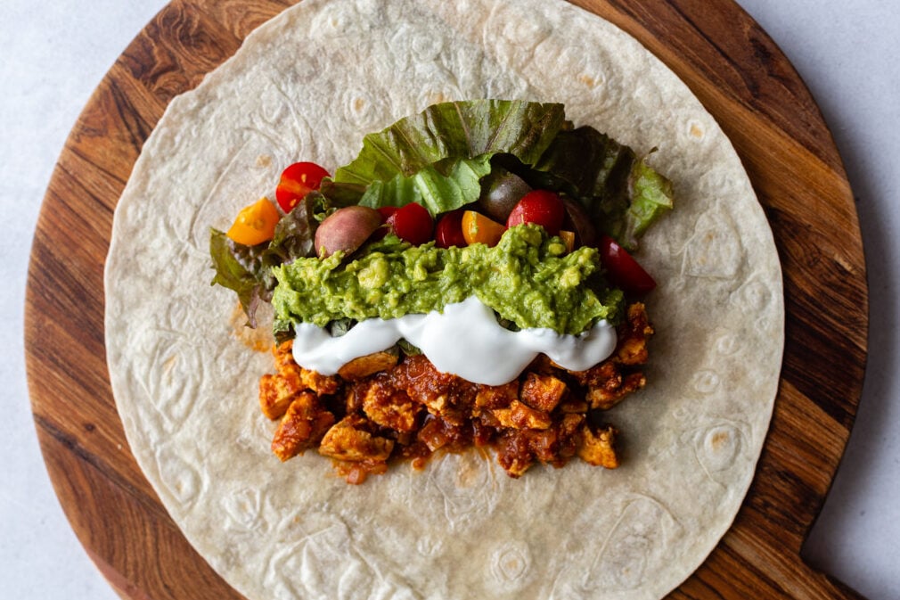 large tortilla sitting on a round wood cutting board. Topped with spicy tofu, sour cream, guacamole, tomatoes, and lettuce