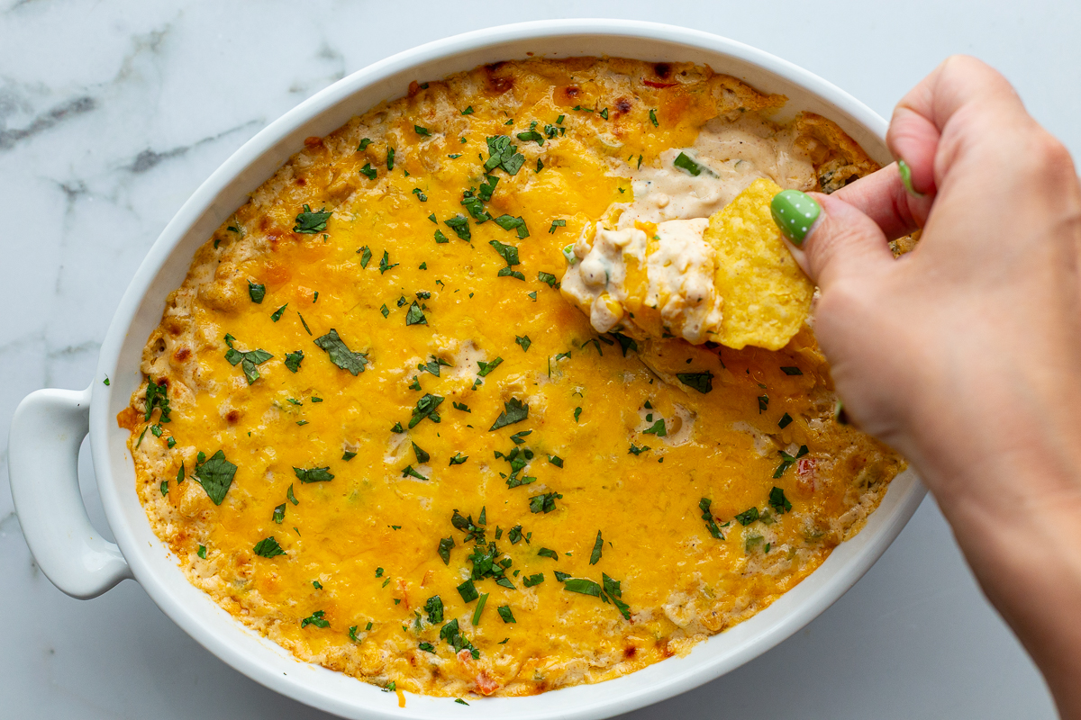 cream cheese corn dip in an oval dish with a hand taking a scoop out with a tortilla chip