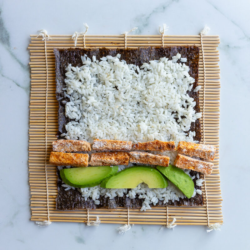rice spread on a nori sheet to make sushi with sweet potato and avocado laying on top before being rolled